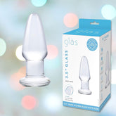 The image displays the Gläs 3.5" Glass Butt Plug along with its accompanying packaging. The predominantly white packaging features light blue accents, the brand name "Gläs," and product details. The background showcases a soft, blurred, colorful bokeh effect, setting an inviting scene for anal play essentials.